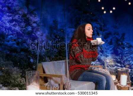 Young woman sitting on a swing with a blanket under the flashlights and holding a cup of coffee in a snow-covered park in the evening, wearing red woolen sweater and knitted scarf while snowing