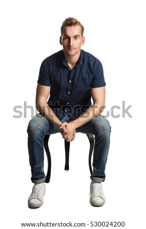 A sad man sitting down on a stool in front of a white background, wearing a blue shirt and jeans with white shoes, looking at camera. Royalty-Free Stock Photo #530024200