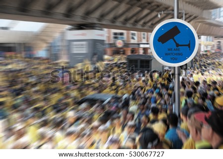 A metal road sign with icon of a closed circuit television and the warning message: We are watching you, over a dynamic view of a street crowd.
