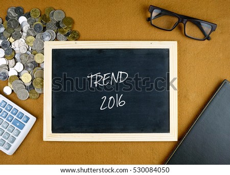 TREND 2016 written on chalkboard, calculator, coins, spectacle and notebook on desk. Flat lay concept.
