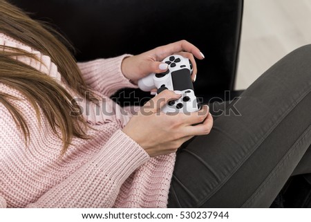 Side view of a woman wearing a pink sweater and black trousers and sitting in a leather armchair with a video game controller