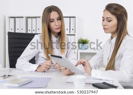 Two women are sitting at a table in an office room. One is working with her tablet while her colleague is watching. Concept of cooperation