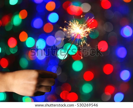 Female hand holding sparkler against colorful defocused lights