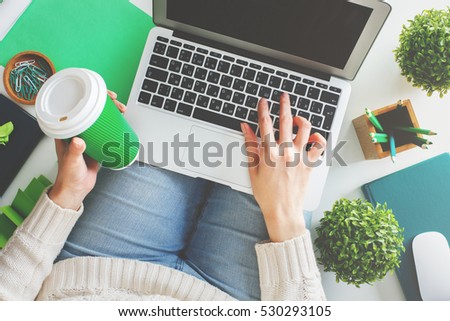 Casual woman holding coffee cup and using laptop with empty screen while sitting on white background with various items. Mock up