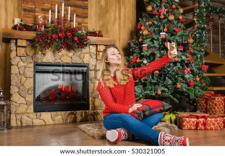 Young woman in country house sitting on the floor, nearly fireplace and give a kiss on mobile. Merry Christmas concept.