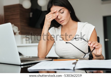 Photo of young tired woman analyzing home finances with laptop while suffering eyestrain and holding glasses. Royalty-Free Stock Photo #530362219