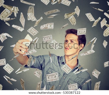 young funny looking man taking pictures of him self with smart phone under money rain  