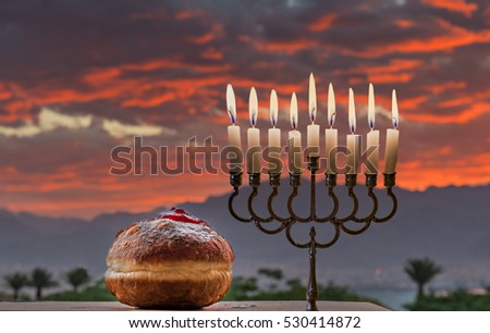 Jewish menorah with the glitter lights of candles and sweet donut are traditional symbols for Hanukkah holiday. Selective focus. Background of colorful cloudscape at dawn taken in Israel