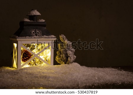 Glowing Christmas lantern casting warm inviting candlelight on winter snow and a small tree with copy space for your holiday greeting