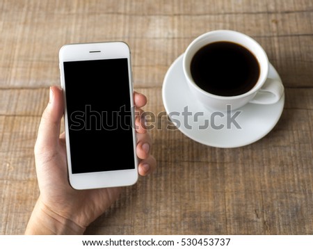 Close up business man's hand holds smart phone with black isolated screen over wooden background and coffee.