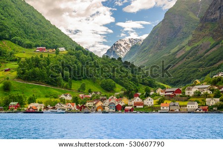 A lots of little colorful traditional Norwegian houses near the sea with green forest on mountains on background and blue sky with dark clouds, Sognefjord, Norway Royalty-Free Stock Photo #530607790