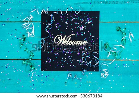 Welcome sign with holiday confetti and ribbon border on blank antique rustic teal blue textured wood background; color copy space for text with white, purple and green decorations
