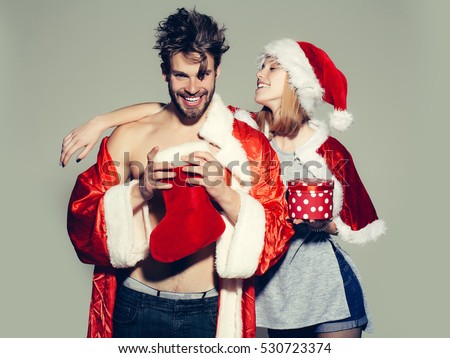 happy young couple of pretty cute girl or woman and handsome bearded man with bare muscular chest in santa claus coat and hat with new year sock or stocking and present or gift box on grey backdrop