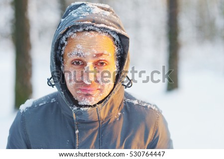 Frozen young man in jacket hood snow covered in winter forest. Snowflakes lie on eyelashes, eyebrows, cheeks frosty face. Bad windy day. Heavy run hard active fit. Strong chill feel, north arctic wind Royalty-Free Stock Photo #530764447