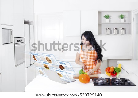 Picture of pretty woman wearing apron while cutting broccoli and looking at a virtual food recipes on laptop in the kitchen