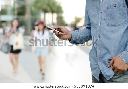 Casual young man in blue shirt and jeans standing and  looking at mobile smartphone at street market with blurred women walking at business area or shopping center  background, communication concept