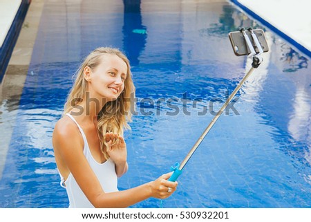 Young blonde caucasian woman in white swimsuit taking selfie picture with smartphone selfie stick next to the pool during luxury vacation in Bali