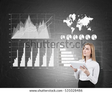 Close up of a blond businesswoman holding a tablet and standing near blackboard with graphs and world map. Elements of this image furnished by NASA