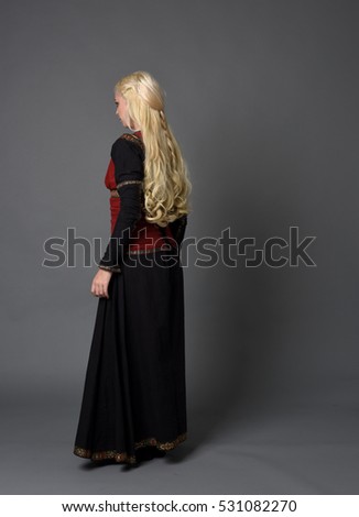 full length portrait of a beautiful blonde woman wearing a black and red medieval gown, standing pose against a grey studio background.