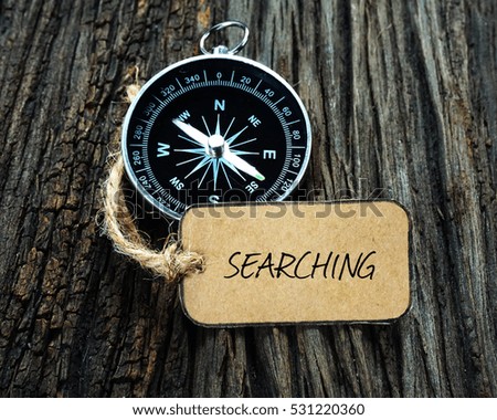 SEARCHING inscription written on paper tag, compass on old wooden background.