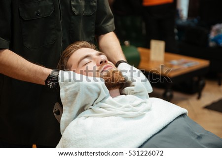 Picture of handsome bearded man getting beard shaving by hairdresser while lies in chair at barbershop.