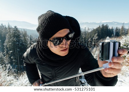 Winter travel. Man in winter wear and sunglasses make selfie using selfie stick in sunny winter weather.