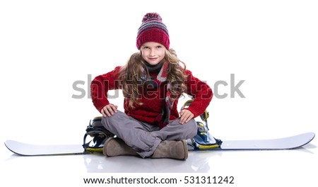 Smiling little girl with curly hairstyle wearing knitted sweater, scarf, hat sitting on blue snowboard, isolated on white. Winter clothes and sport concept.