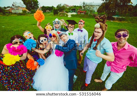 Bride in funny glasses and groom in wolf hat pose with their masked friends on the field
