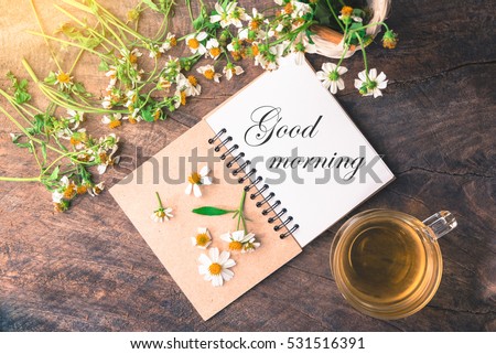Good morning text on notebook with white flower and bas ket of flower and cup of tea on vintage wooden table View from above with warm light.