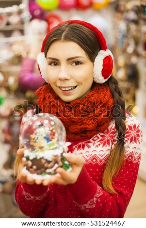Brunette girl in earmuffs buy present at mall.Shopping center decoration.Pretty white woman shopper buy New Year presents for friends and family in store.Holiday decorated shop.Female hold snow sphere