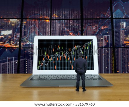 Businessman standing on the wood table which have the computer laptop showing the trading graph at the screen over the blurred photo of trading graph on cityscape background, Business trading concept