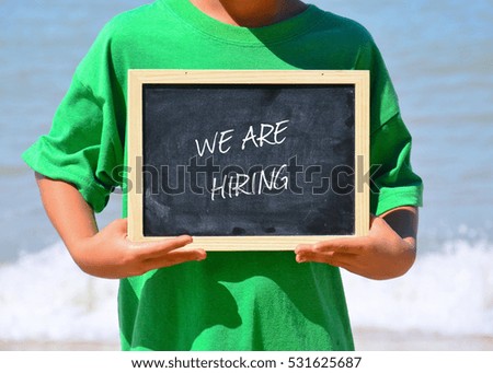 A man holding a chalkboard written with WE ARE HIRING text. Wave and blue ocean background.