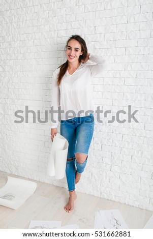 Beautiful barefoot woman smiling at camera. Full-length portrait of attractive female artist with sketch leaning on white brick wall at workshop. Art, creativity, relax concept