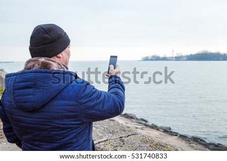 man in a cap and a blue jacket photographing seascape by mobile phone on cold autumn day
