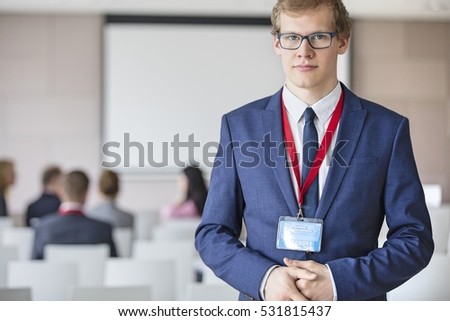 Portrait of confident businessman standing at seminar hall with colleagues sitting in background