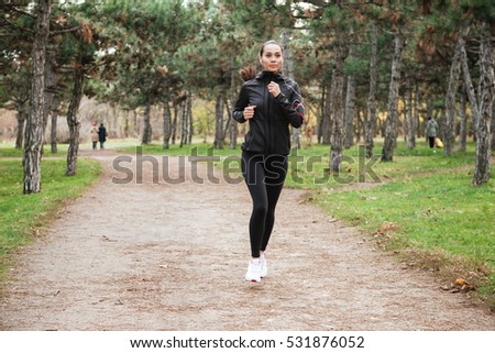 Picture of young cheerful lady runner in warm clothes and earphones running in autumn park