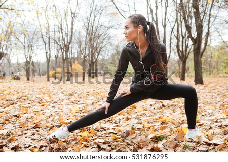 Picture of attractive woman runner in warm clothes and headphones looking aside in autumn park while make sport exercise