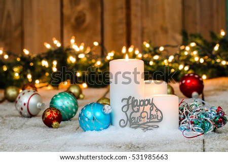 White holiday candles, Happy New Year and colorful ornaments and ribbon on snowy rustic wooden background with garland and string of lights border; holiday background with white copy space