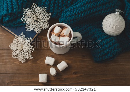  cozy winter home background - cup of hot cocoa with marshmallow, Christmas ball, snowflakes and warm blue knitted sweater, book on wooden table.