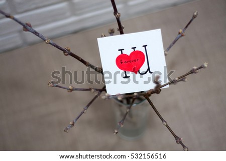 Gray love background with red heart and written saying wise quote "I love you forever" on a piece of paper card placed on branches in a vase on linen cover on the table in room with white brick wall