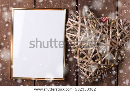 Empty golden frame and decorative heart with fairy light  on  aged wooden background. Place for text. Mock-up for design. Drawn snow.