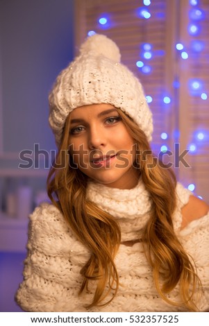 Very beautiful young girl with long brown hair in white hat and a white scarf on a background of blue Christmas lights