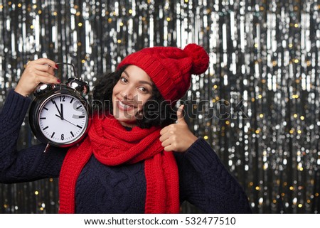 New Year eve concept. Happy woman in winter hat and scarf holding with big alarm clock counting to midnight and showing approving thumb up gesture
