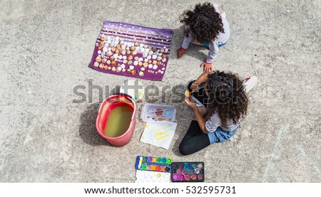 Brazilian girls painting shells outside on a summer day. Top view at two kids sitting on a patio floor painting with watercolors, image for creative family concept, hobby blog, toys  business website