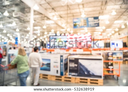 Blurred TVs shopping at large wholesale club. Television retail shop, row of big screen, smart TVs display on shelves. Customers browsing to select and use flatbed cart to carry TV to checkout counter