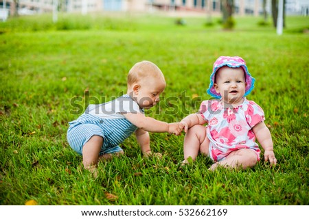 Two happy baby boy and a girl age 9 months old, sitting on the grass and interact, talk, look at each other. Kids are resting on a hot summer day on the grass.