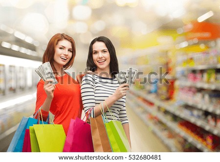 sale, finances and people concept - two smiling teenage girls with shopping bags and cash money over supermarket background