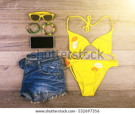 Set of beach clothes yellow bikini, bracelets, jeans shorts, glasses on dark wooden background. Top view. Summer Holiday Concept.
