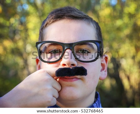 a young boy holding a finger mustache up to his face wearing a pair of urban hipster glasses and making a serious face in a sunny outdoor setting 