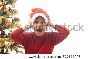 Schoolboy dressed in red with a santa hat isolated on white background. Christmas tree in the background. Happy child covering his ears. Space left for copy